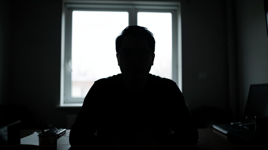 Une personne est en silhouette devant une fenêtre lumineuse dans une pièce sombre, avec un bureau et du matériel informatique visible.